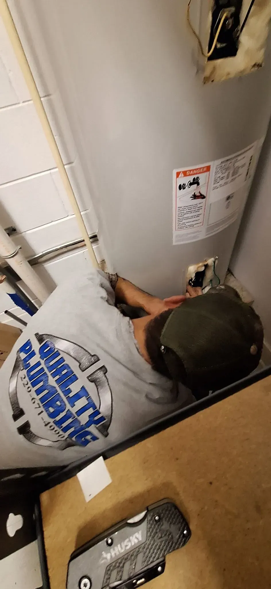 A plumber works on a water heater. He wears a Quality Plumbing shirt and a cap, with a utility knife in front.