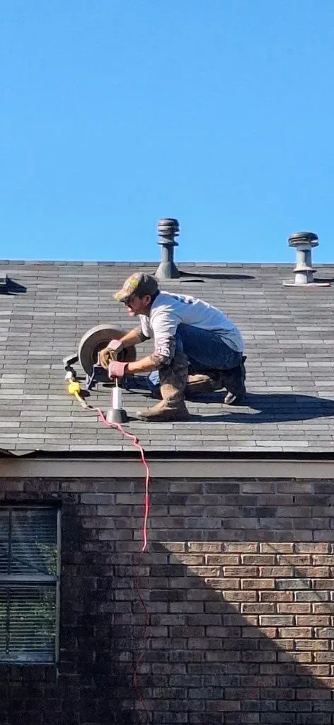 Man on roof using a tool, with a red rope. Brick building and blue sky background.