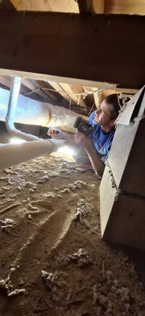 A person examines something in a crawl space, using a flashlight. Insulation and ductwork visible.