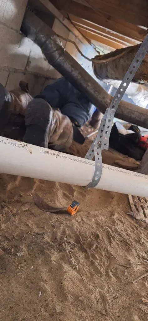 Person working under a building, visible legs, white pipe overhead, dirt ground, and a small tool.