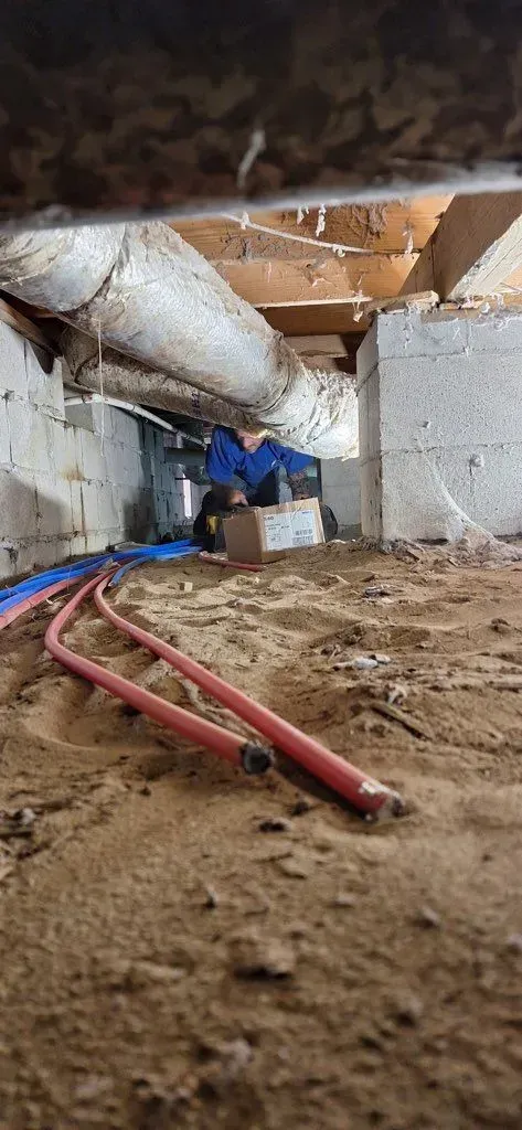 An crawlspace with insulation, wood beams, and red piping on a sandy floor.