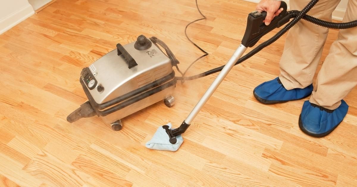 Person steam-cleans a wooden floor with a white cleaner and gray machine while wearing blue shoe covers.