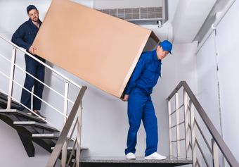 Two men are carrying a large cardboard box up a set of stairs.