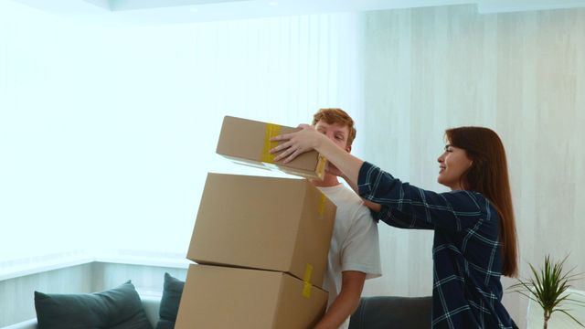 A man and a woman are carrying boxes in a living room.