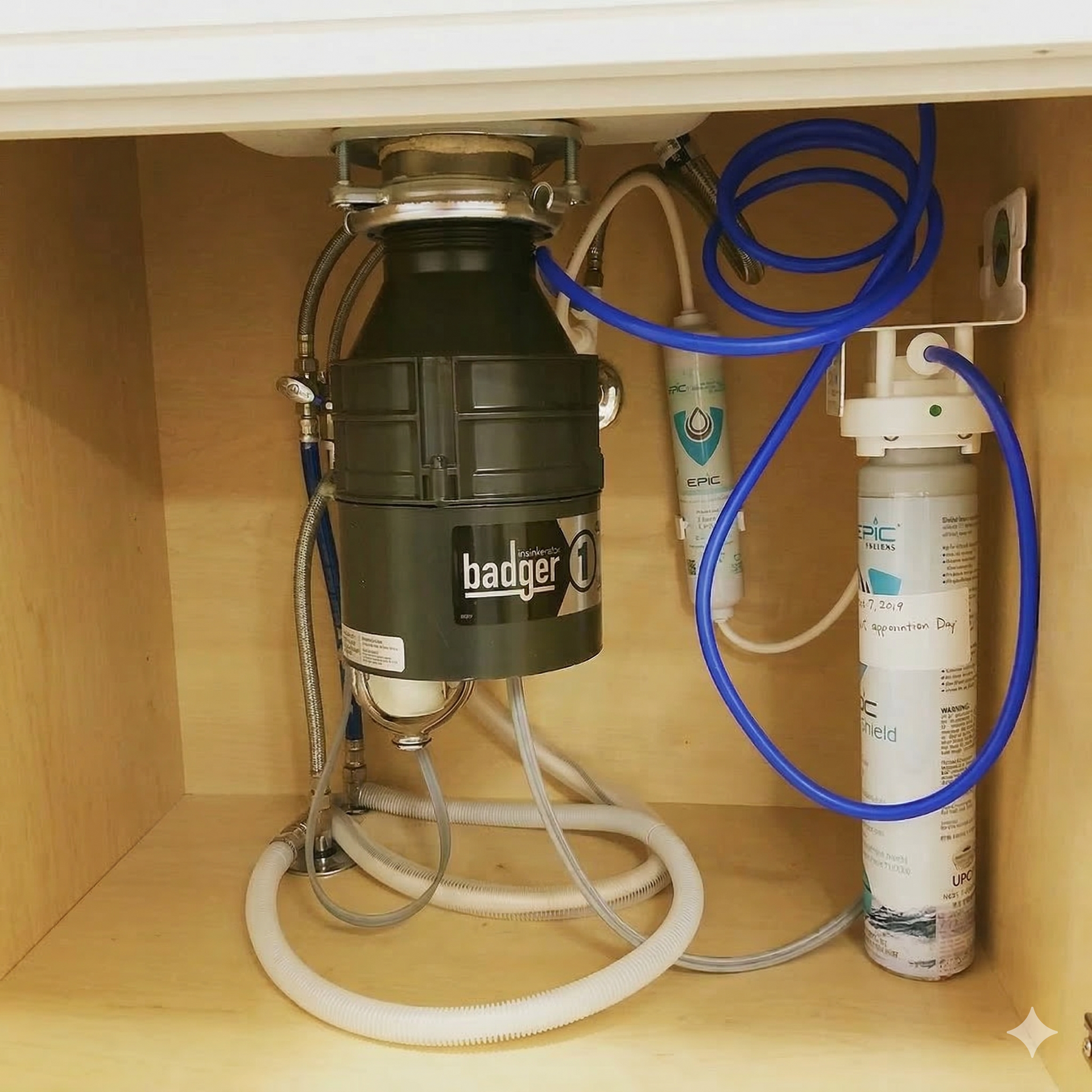 A kitchen cabinet interior containing a Badger garbage disposal unit and a water filtration system with blue tubing.