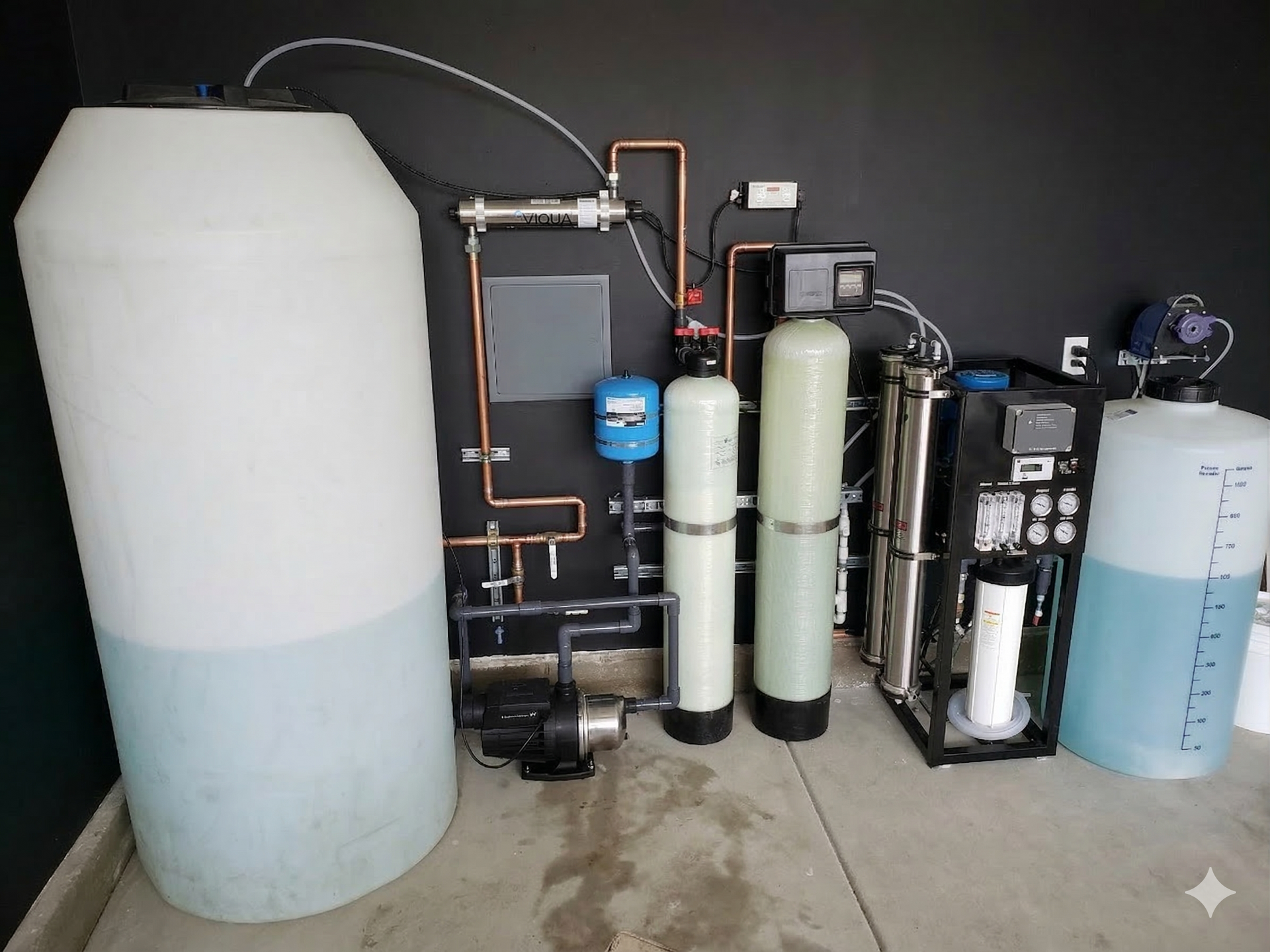 A residential water treatment system featuring tanks, filtration columns, a pump, and piping against a black wall.