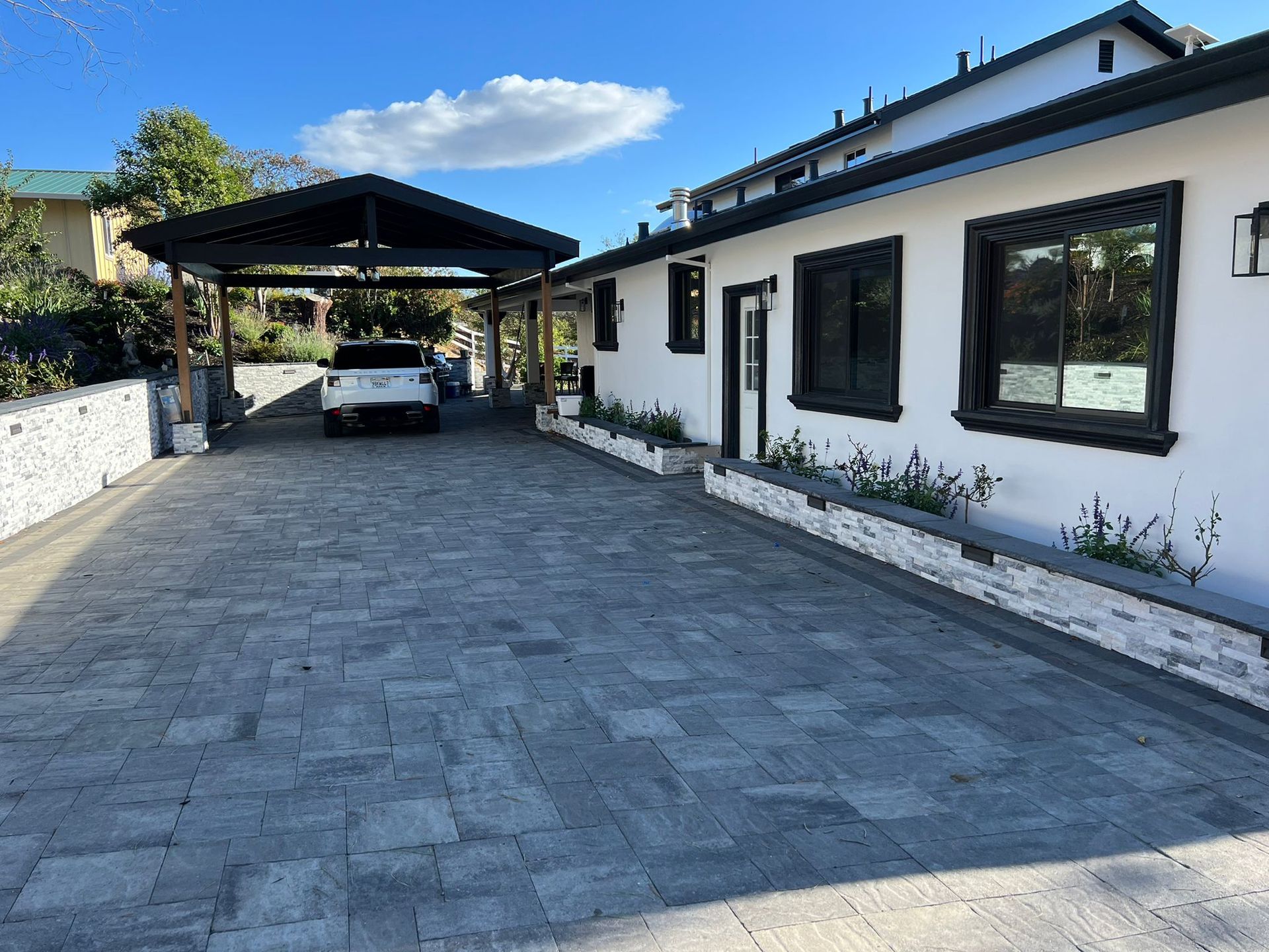 A white car is parked under a carport in front of a white house.