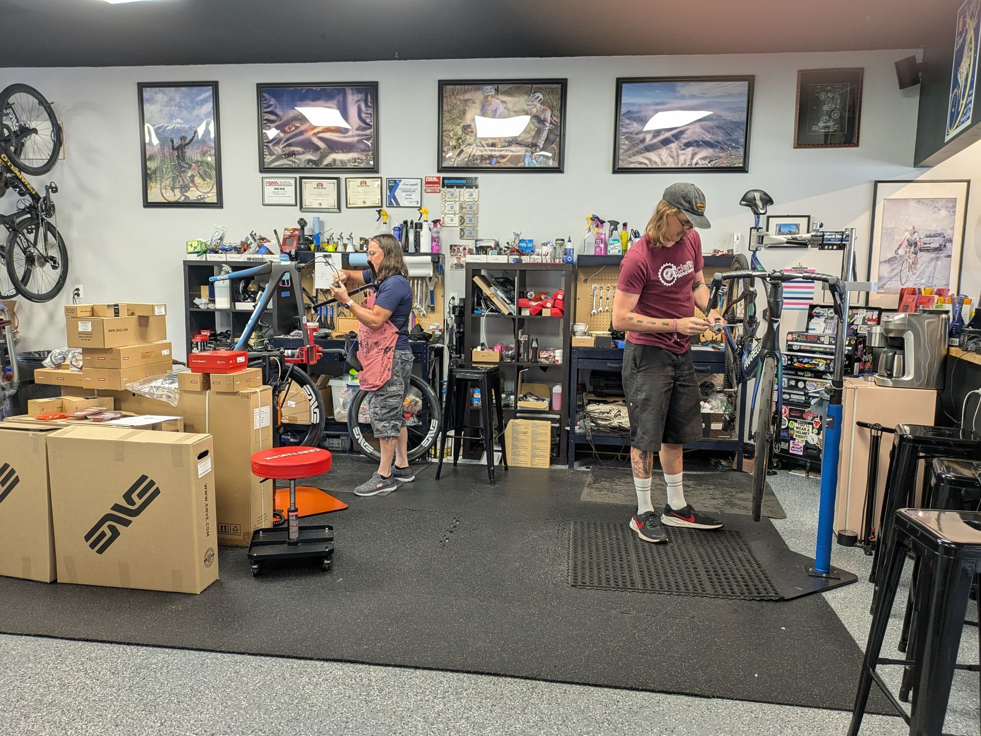 Two people working on bikes in a bike shop; boxes, tools, and art on the walls.