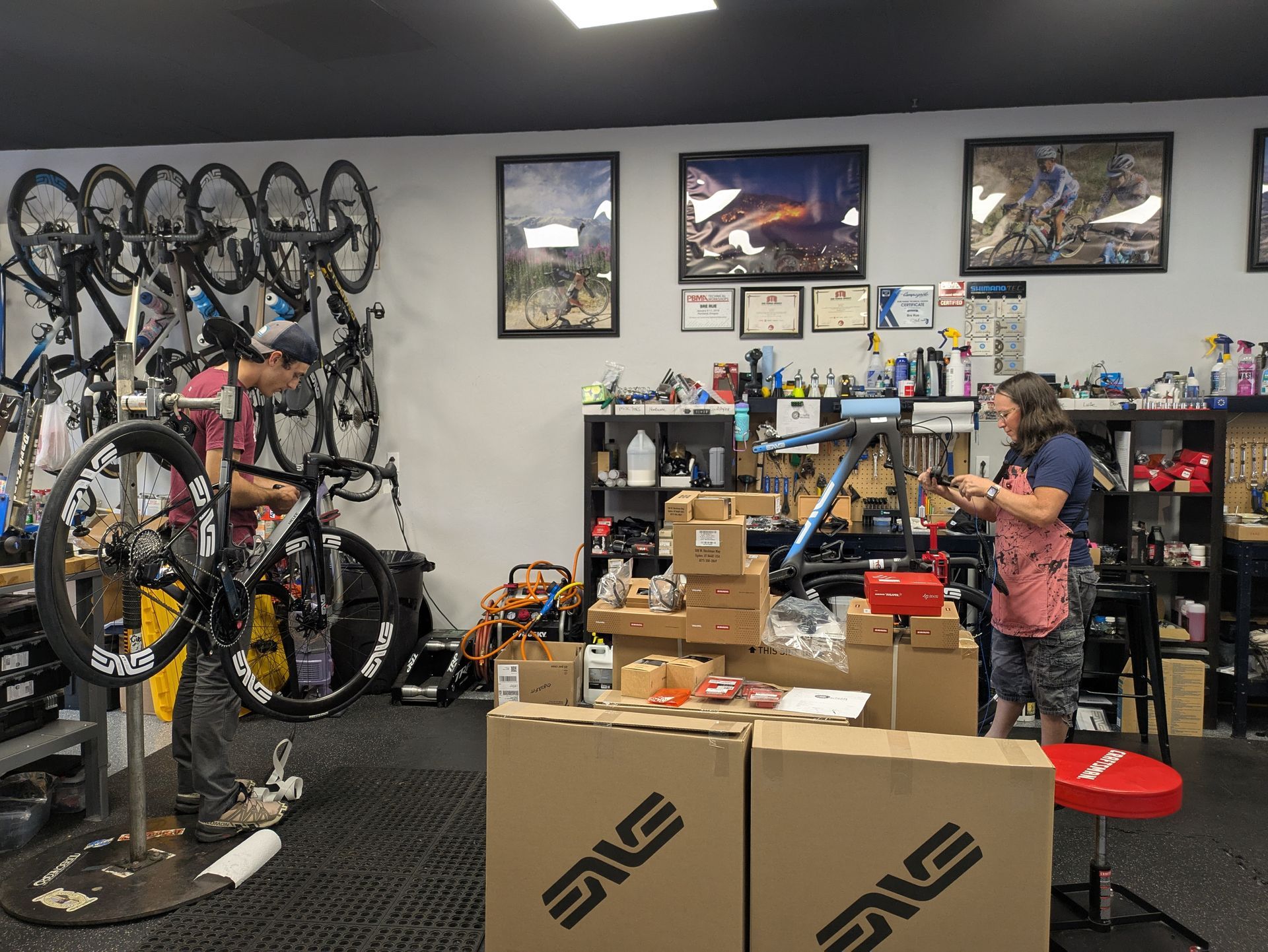 Two people in a bike shop. One repairs a bike, the other organizes parts. Bikes and supplies surround them.