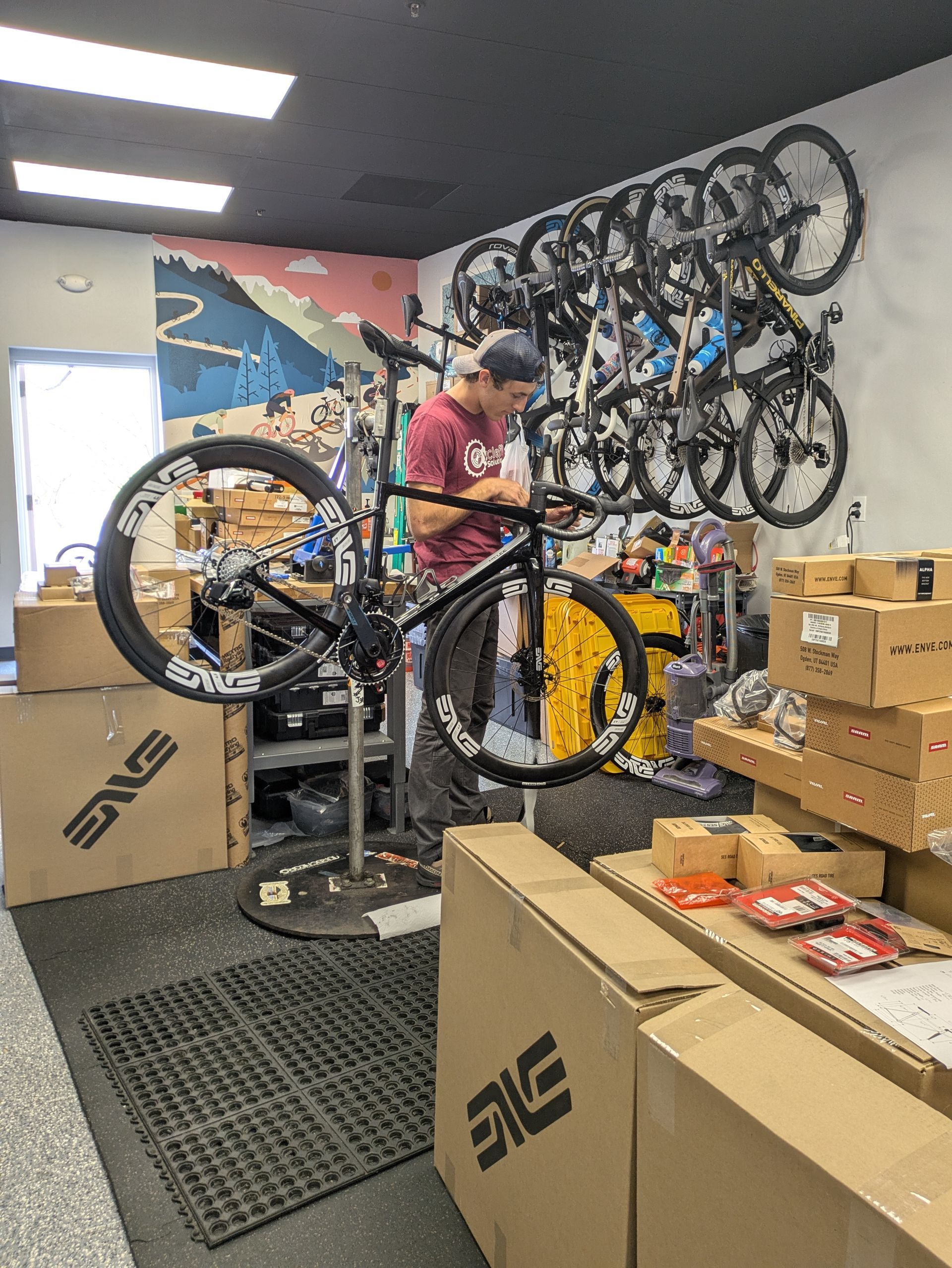 A person working on a bicycle in a bike shop. Several bikes on wall. Boxes and equipment around.