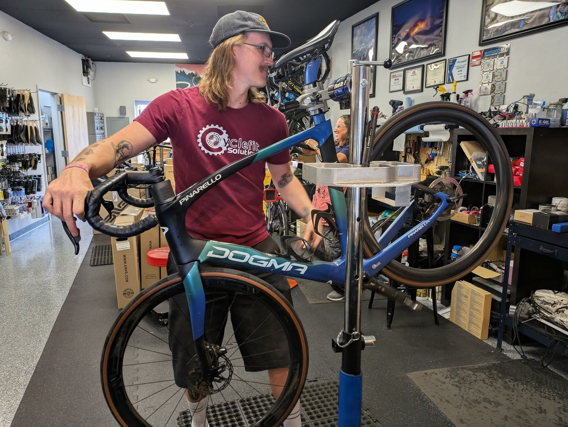 Man working on a blue and teal bicycle in a bike shop. He wears a red shirt and a baseball cap.