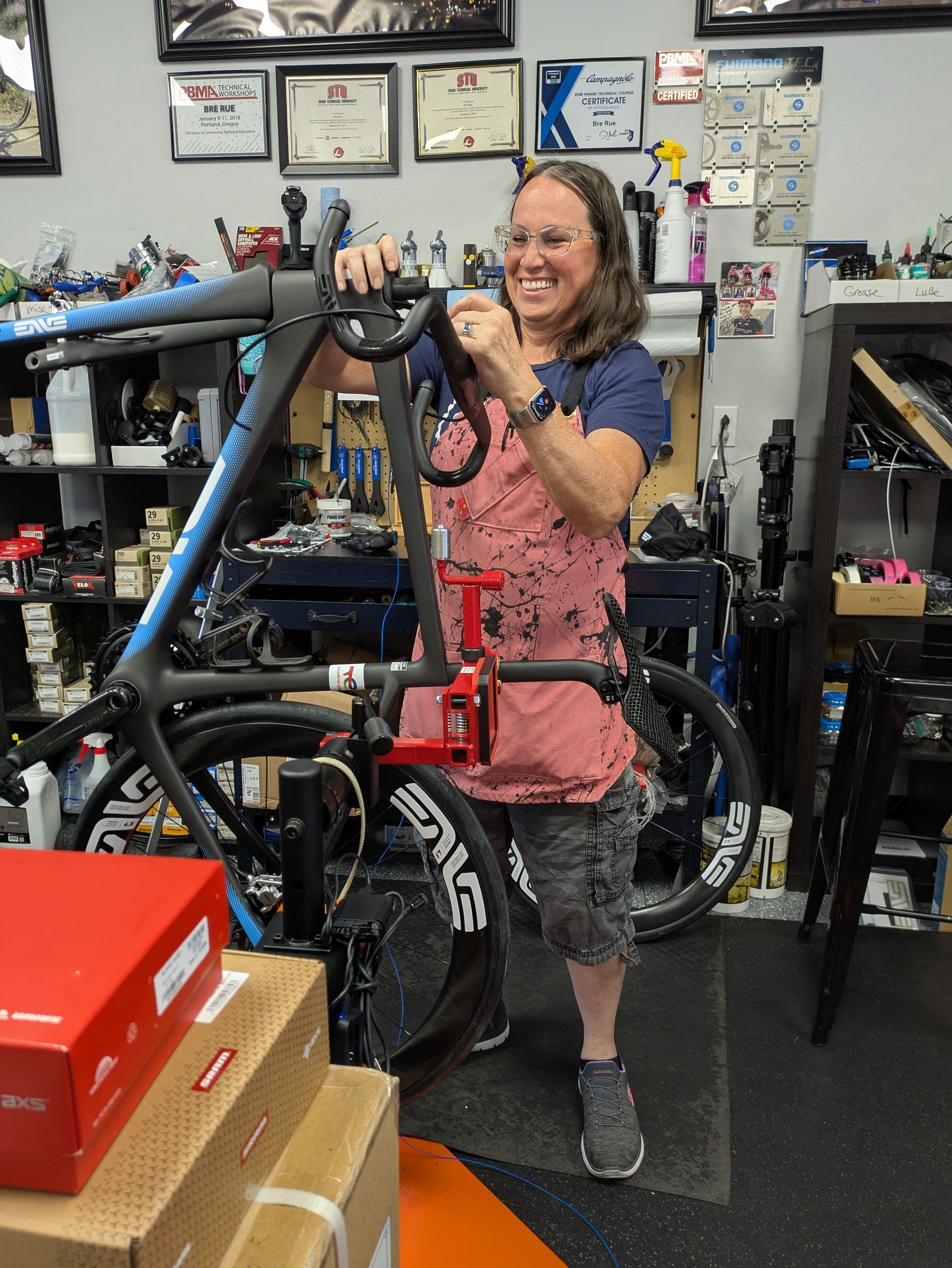 Woman holding bicycle handlebars in a bike shop, smiling. Red work stand supports the bike. Shop cluttered with equipment.