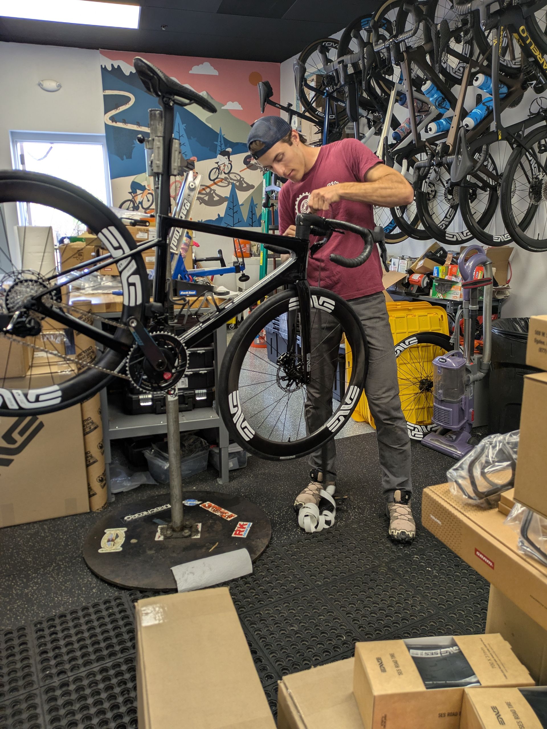 Man works on a black bicycle inside a bike shop. He wears a red shirt and a cap, standing near tools.