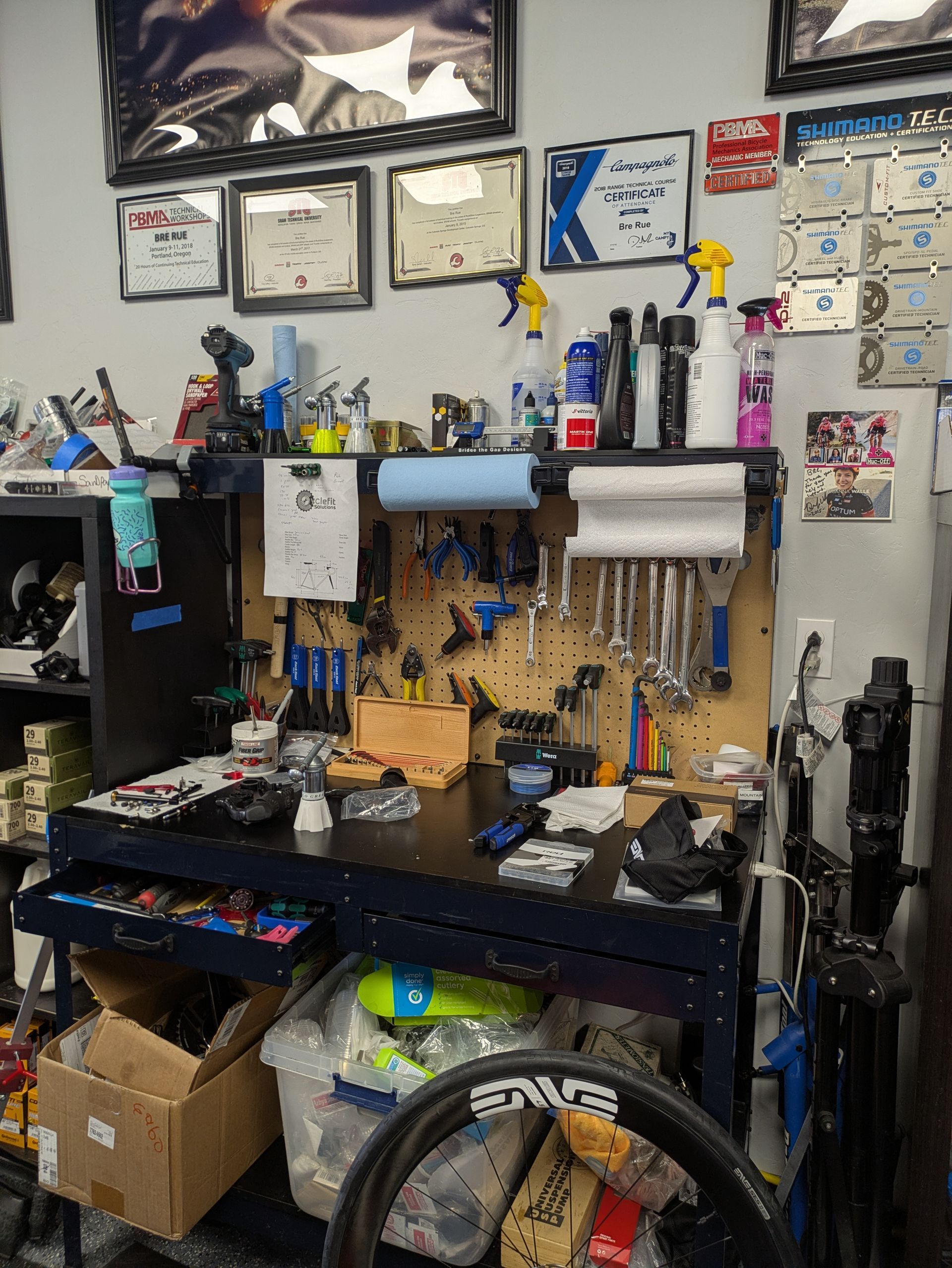 Work bench in a bike shop with tools, parts, and a wheel. Certificates and spray bottles on the wall.