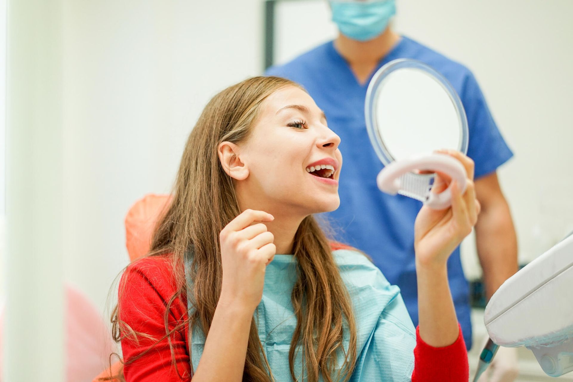 Woman in dental chair looking in a mirror at her teeth with a dentist in the background.