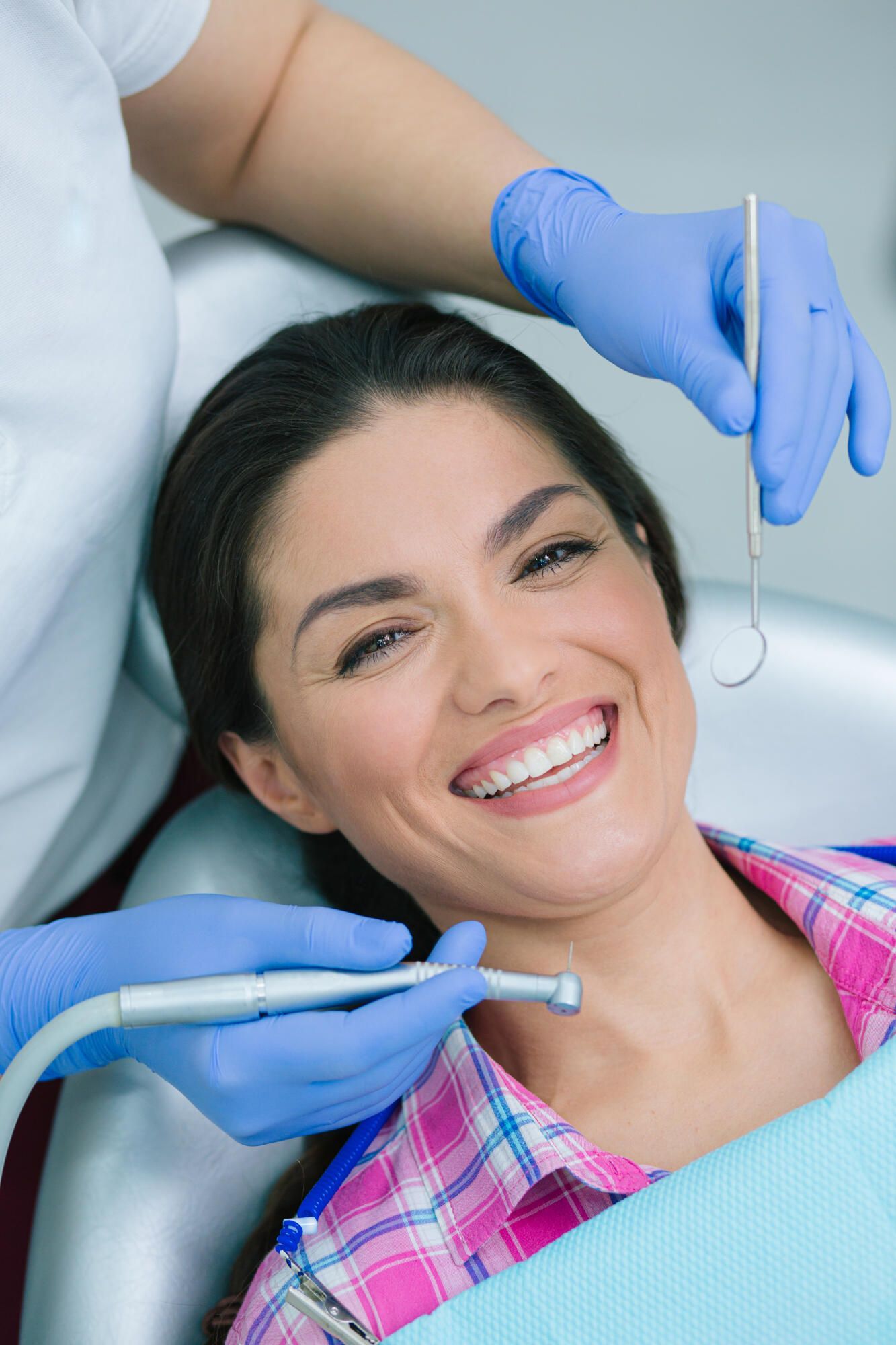 A dentist in blue gloves holds tools while examining a smiling person in a dental chair.