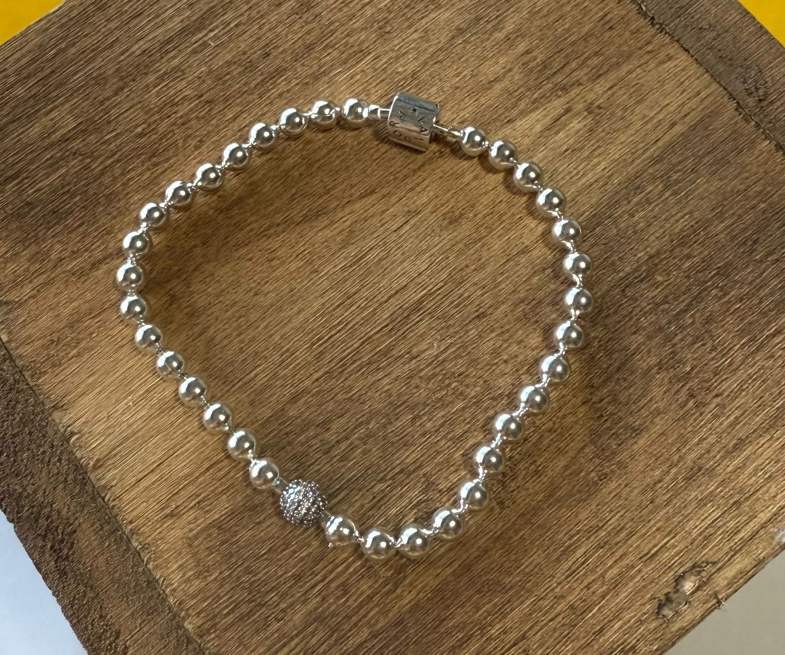 Pulsera de cuentas de plata sobre superficie de madera, con cierre cuadrado y detalle de cristal.