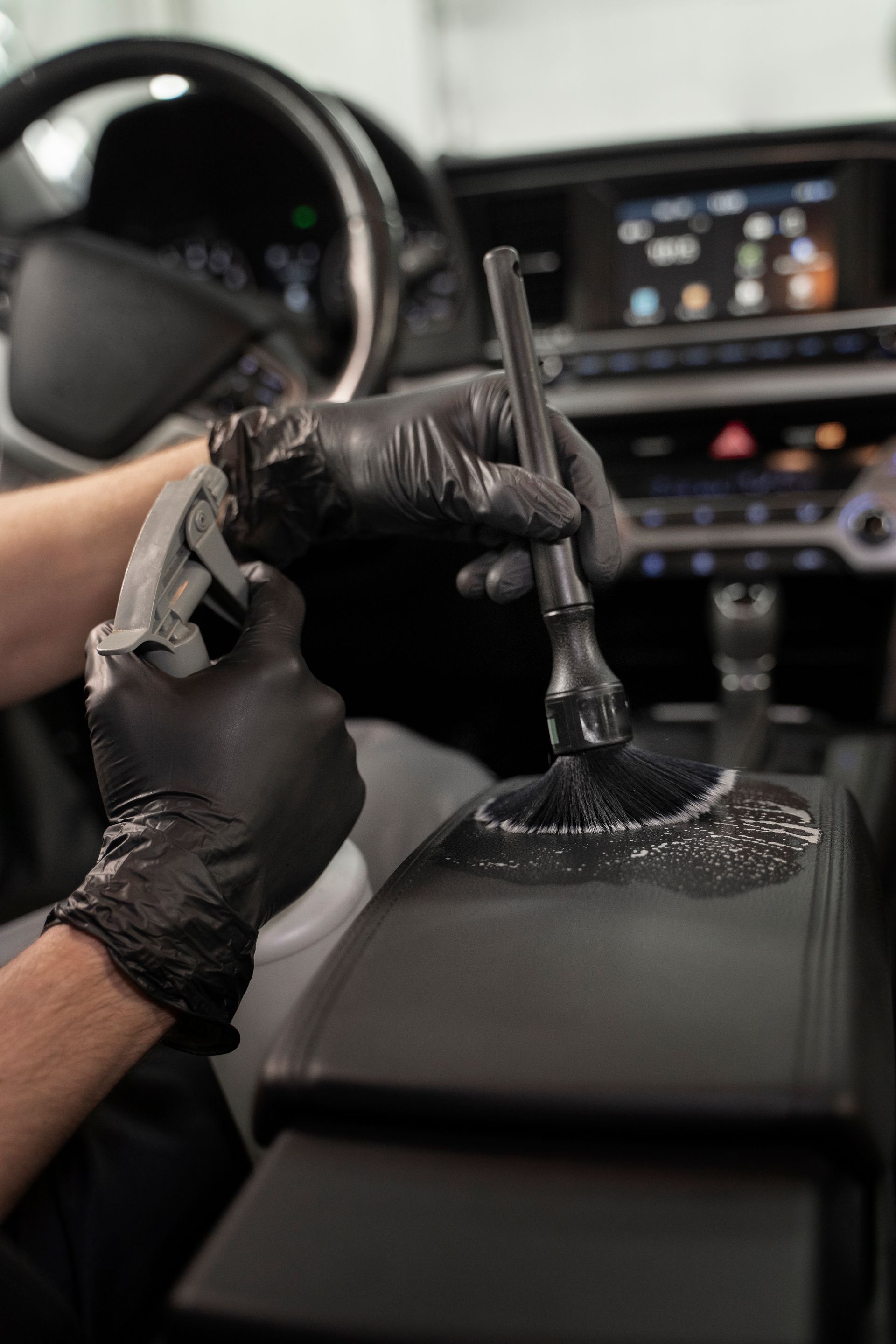 Person wearing black gloves cleaning a car's center console with a brush and spray bottle.