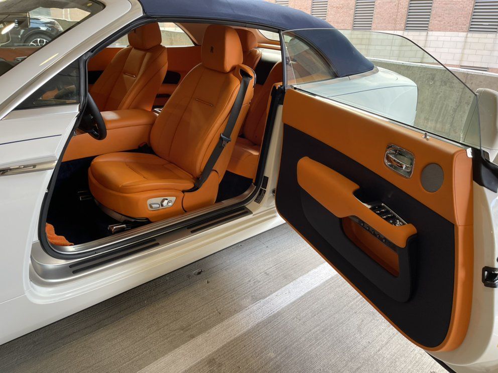 White Rolls-Royce convertible's interior, with tan leather seats and open door. Blue roof, parked indoors.