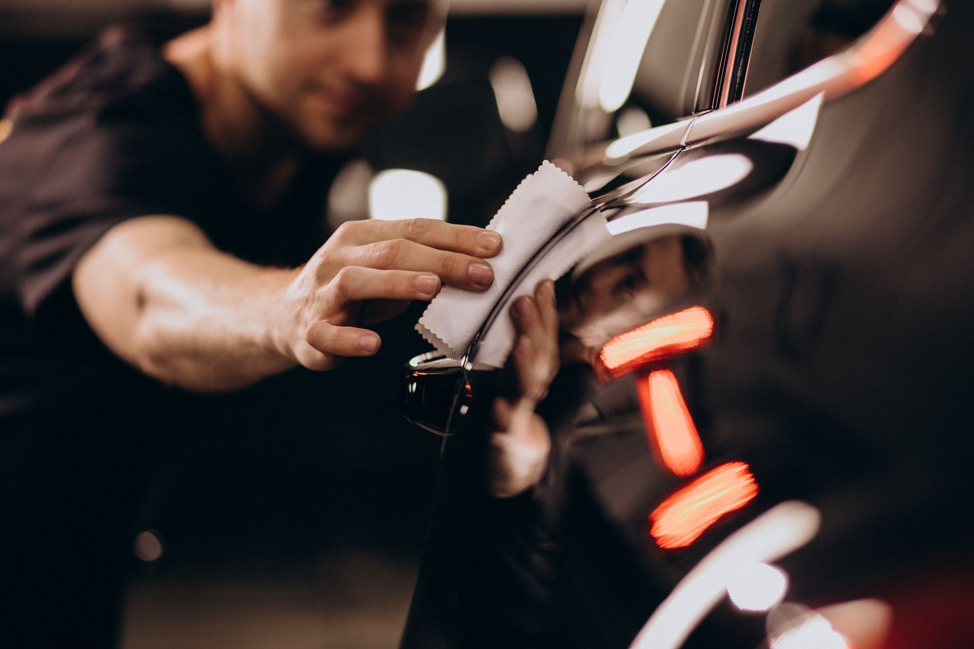 Man polishing a car's chrome detail with a white cloth; blurred background.