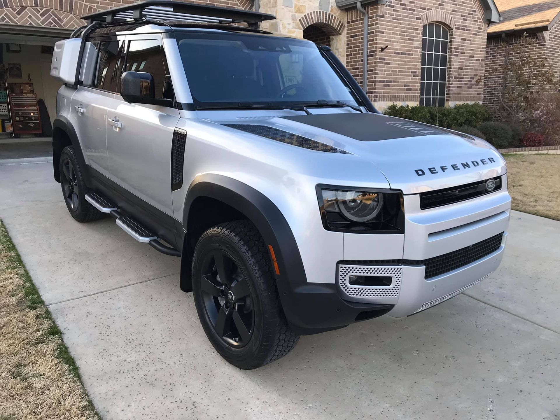 Silver Land Rover Defender SUV parked on a driveway with black accents, roof rack, and a storage box.