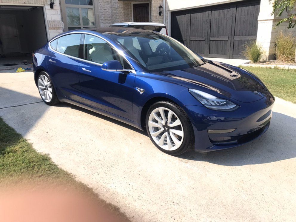 Blue Tesla Model 3 parked on a driveway. Shiny blue car, white wheels, black roof, sunny day.