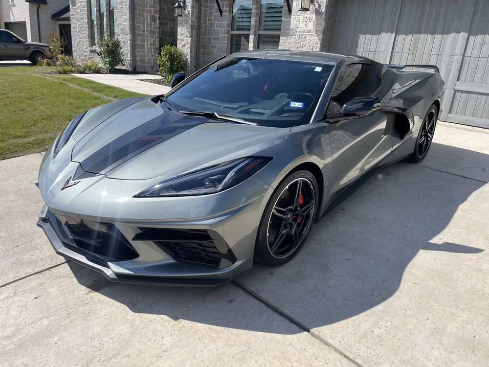 Gray Corvette sports car parked in front of a house with a driveway. Black racing stripe on the hood.
