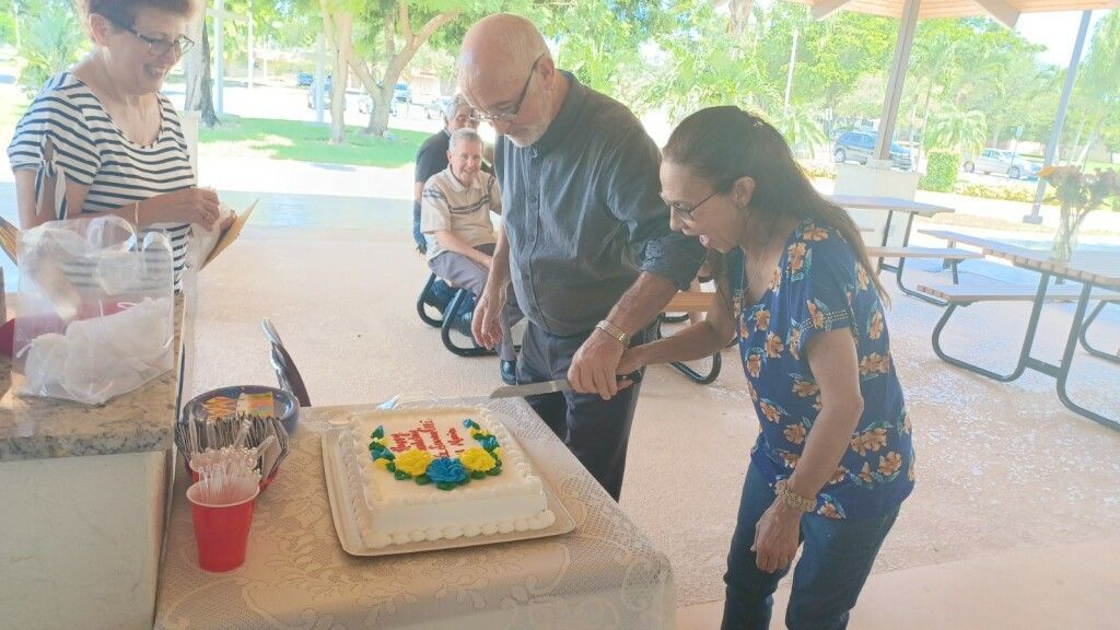 People gathered around a table as a man and girl cut a decorated cake under a park pavilion.