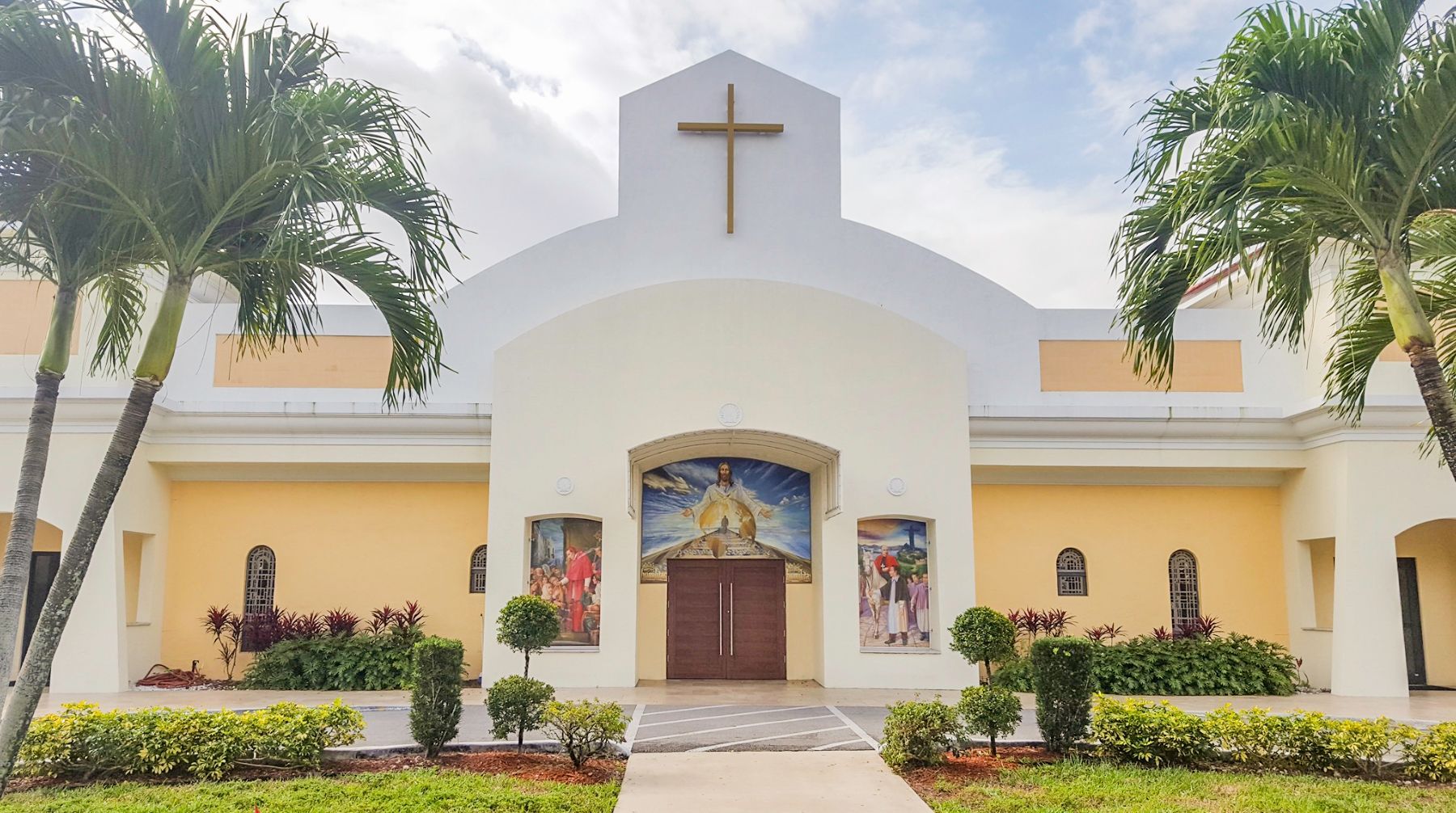 White church facade with a cross, arched entrance, and palm trees in a landscaped courtyard.
