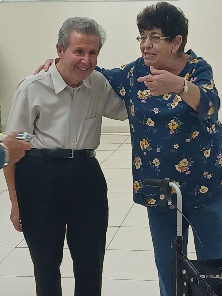 Two people smiling and posing with arms around each other indoors; one uses a walker.