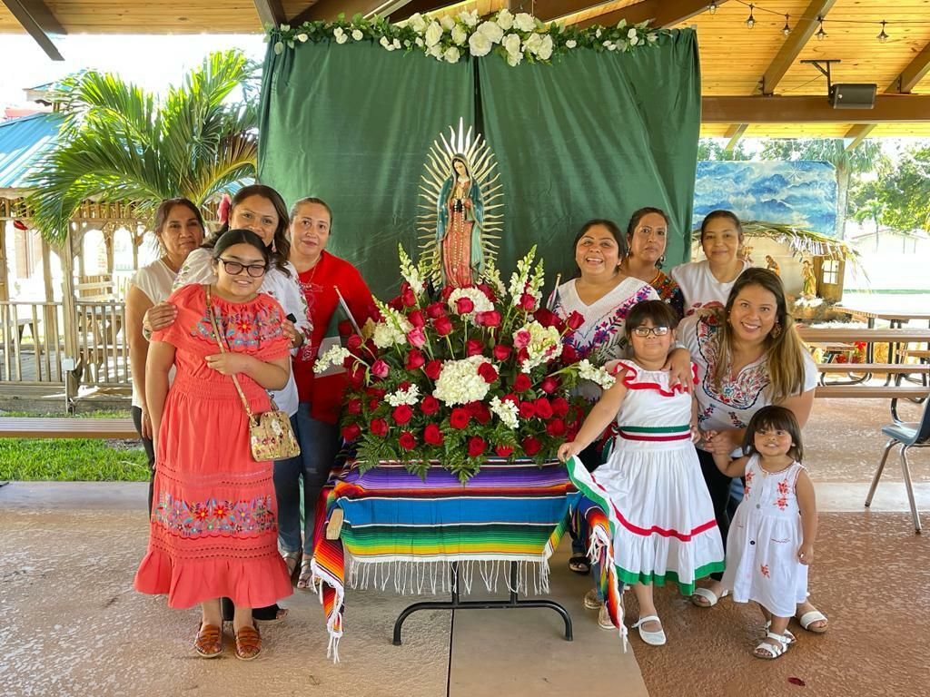 Group gathered around a flower altar with a religious statue under a pavilion, smiling for a ceremony photo