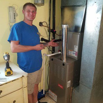 Man in blue shirt and shorts stands by a furnace, tools on a counter, in a basement.