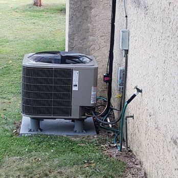 Air conditioner unit on a concrete pad next to a stucco building, with a water hose and electrical box.