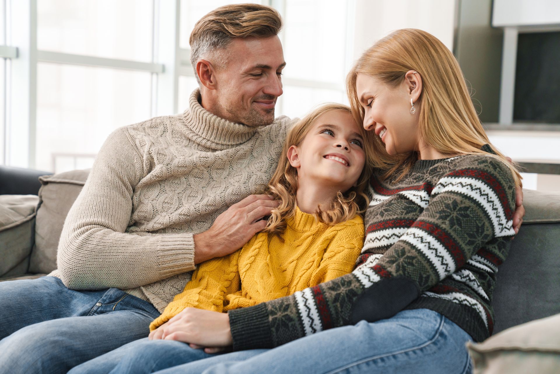 Family of three, seated on a sofa indoors, smiling and embracing.