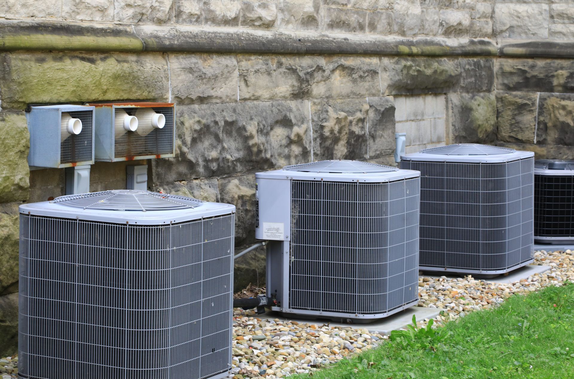 Three outdoor HVAC units installed on gravel near a stone wall for building climate control
