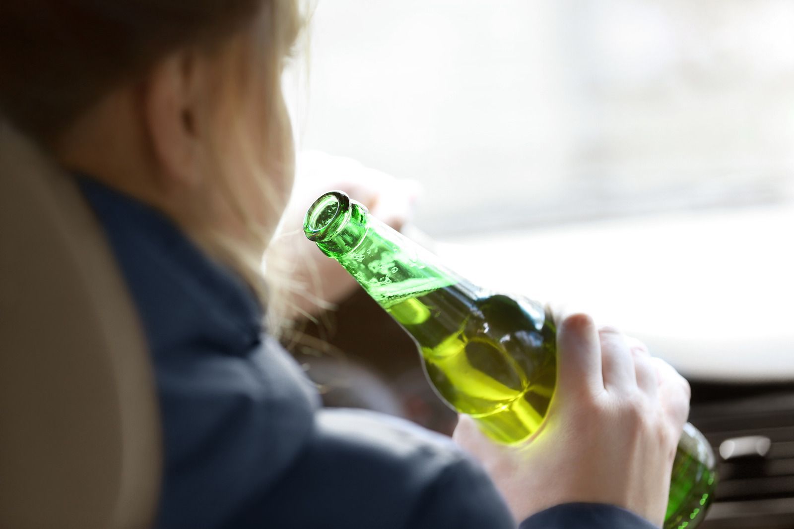A woman is sitting in a car holding a green bottle of beer.