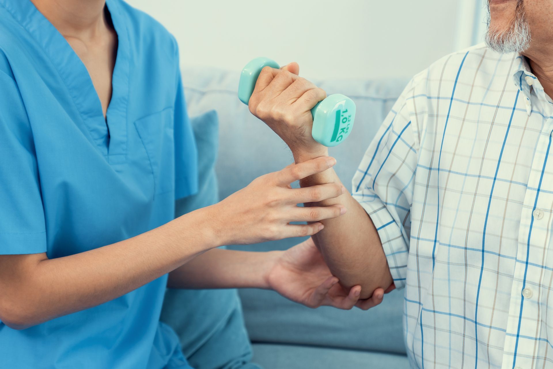 A healthcare worker assists an elderly man with arm exercises using a small weight. They are indoors.