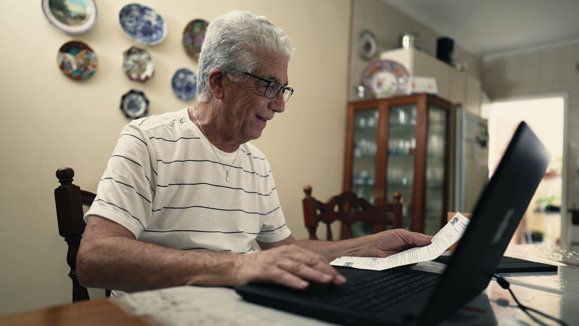 Older man with gray hair and glasses uses a laptop while holding a receipt at a table in a dining room.