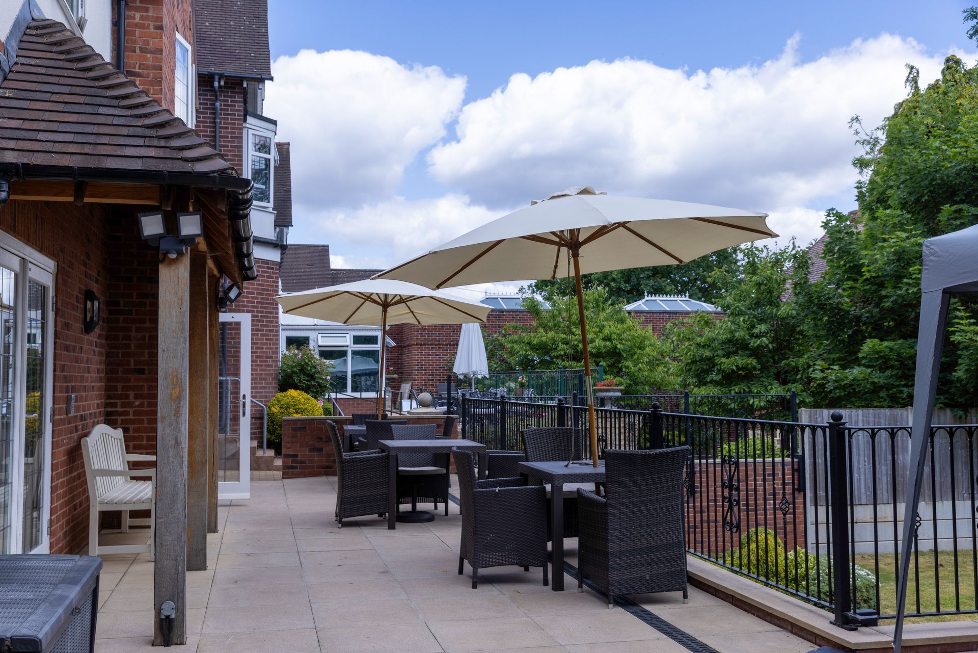 Outdoor patio with tables, chairs, and umbrellas on a sunny day. A brick building and trees are in the background.