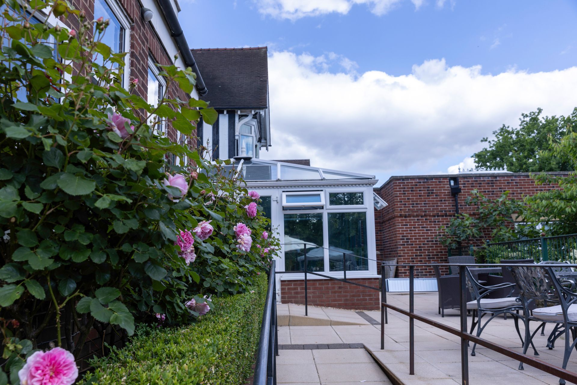 Pink roses bloom beside a brick building with a patio and tables. A conservatory and cloudy sky are in the background.