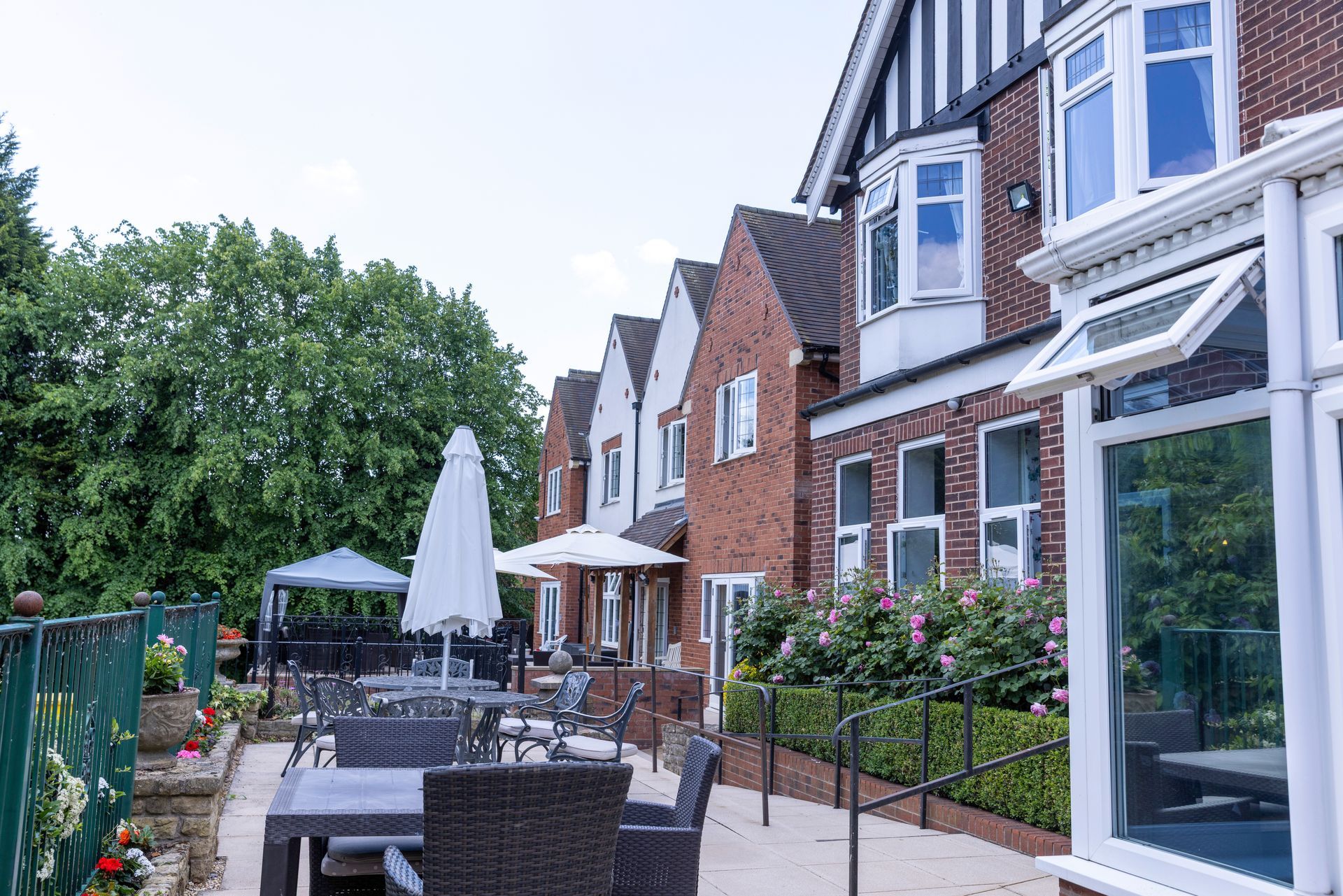 Exterior view of a brick building with outdoor seating area; tables, chairs, and a parasol. Bushes and greenery surround.