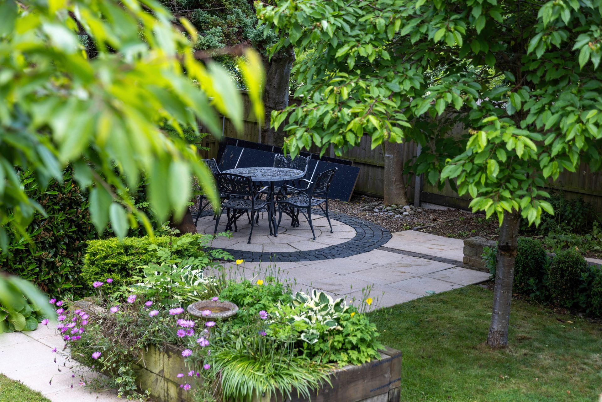 A garden patio with a table and chairs under a tree, surrounded by lush greenery and flowers.