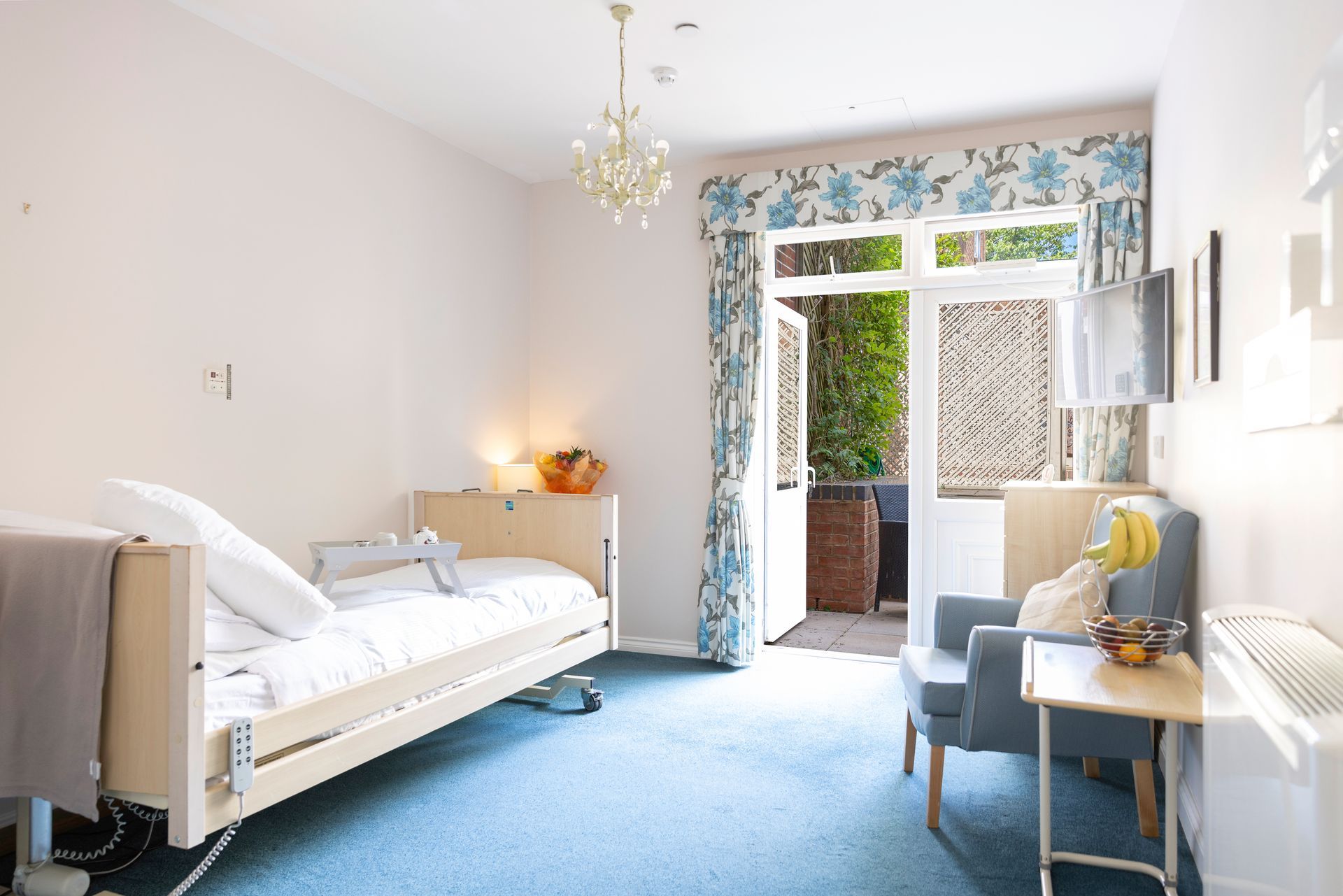 A light-filled bedroom in a care home with a single bed, armchair, and access to a small patio.
