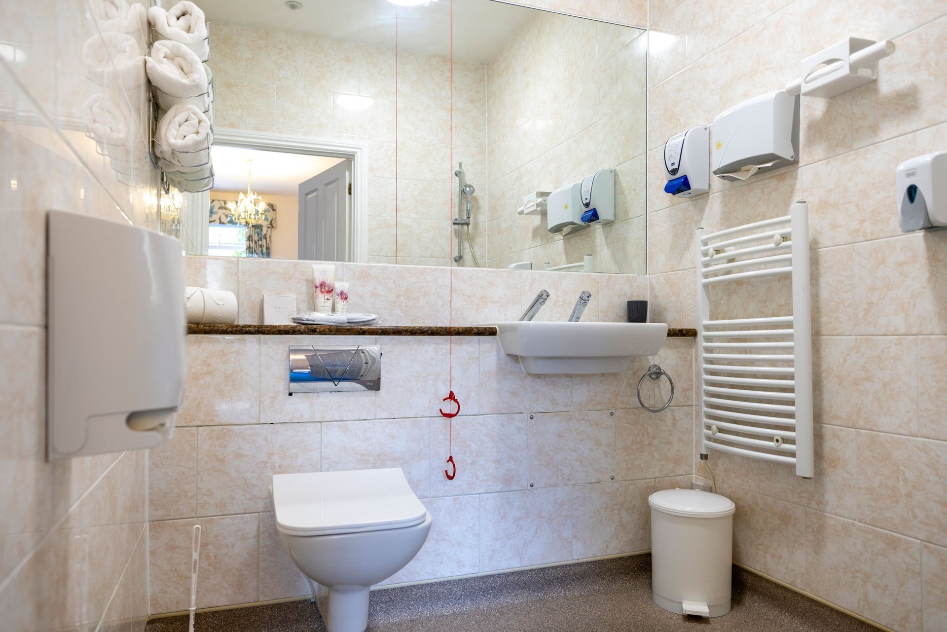 Accessible bathroom with toilet, sink, grab bars, and towel rack; light beige tile.
