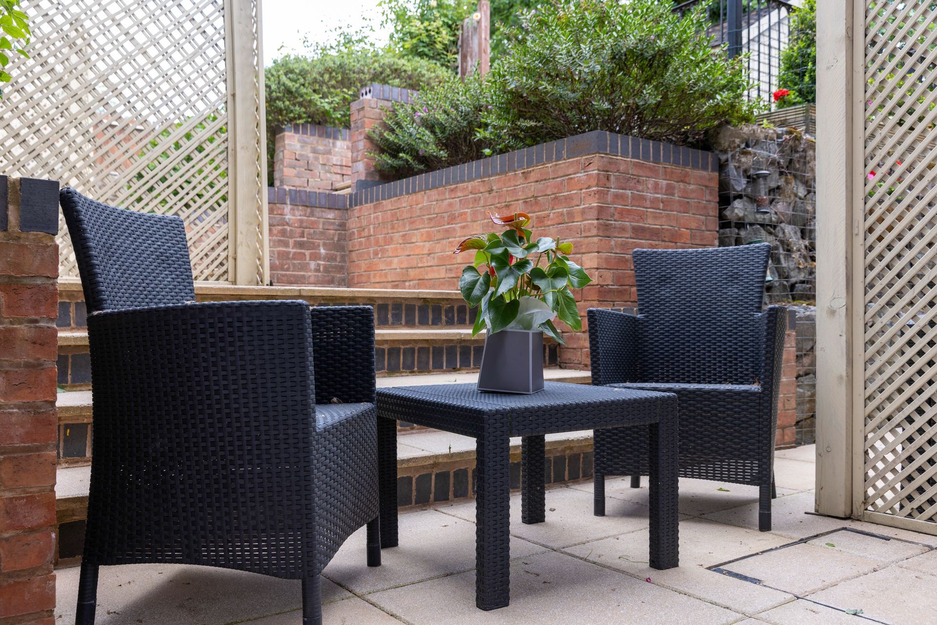 Two black wicker chairs and a small table on a patio with steps and a brick wall, greenery in the background.