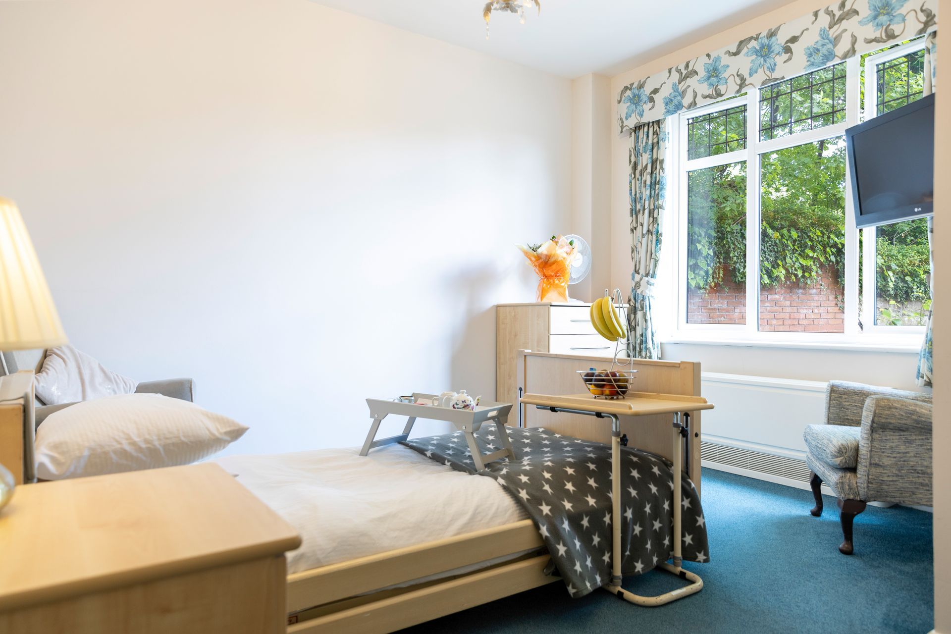 Bedroom with hospital bed, side table, armchairs, window with floral valance, and TV.