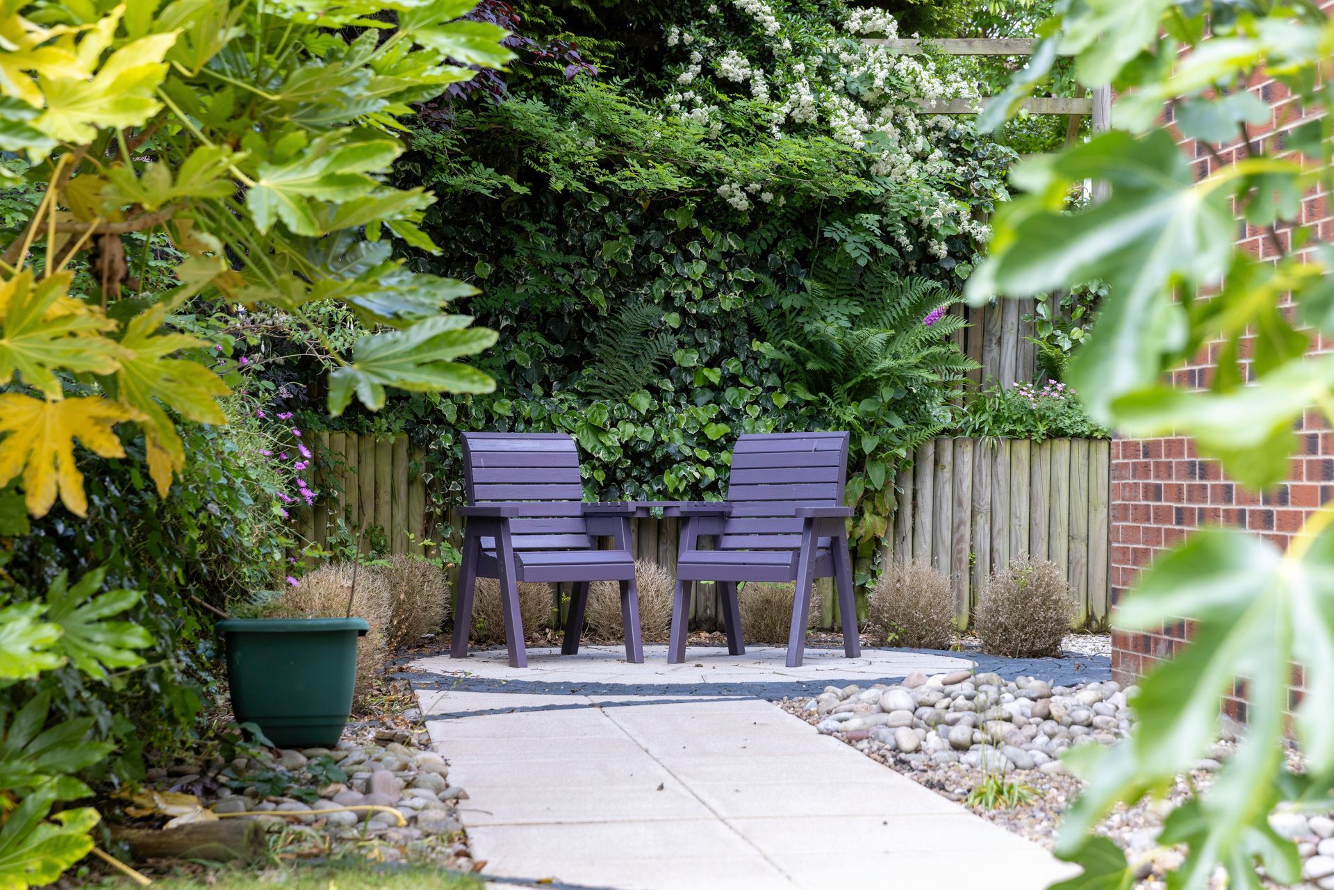 Two purple chairs and a small table on a patio in a garden setting. Lush greenery frames the space.