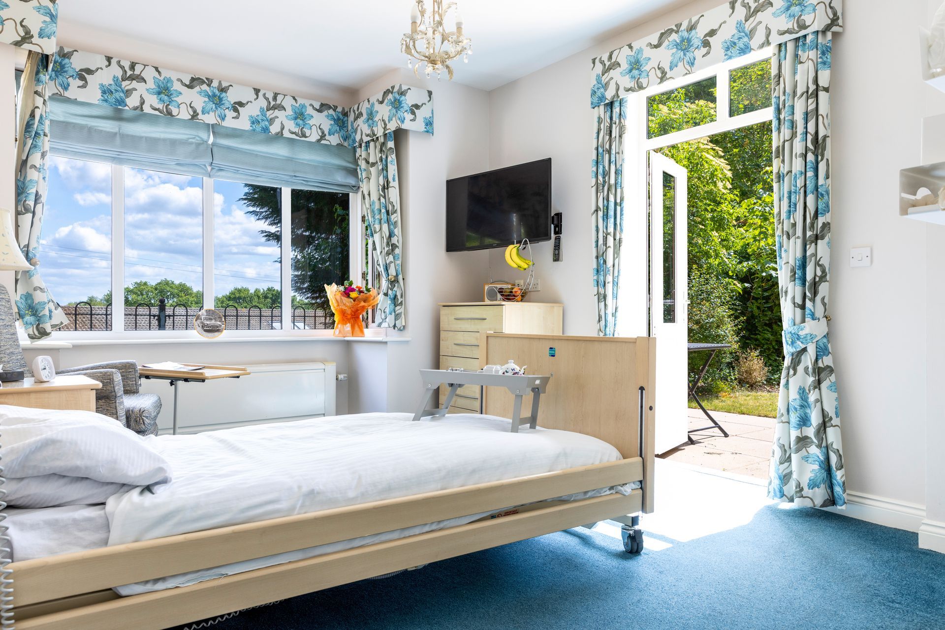 Bedroom with a bed, open doors to a garden, and floral curtains. Blue carpet and white walls.