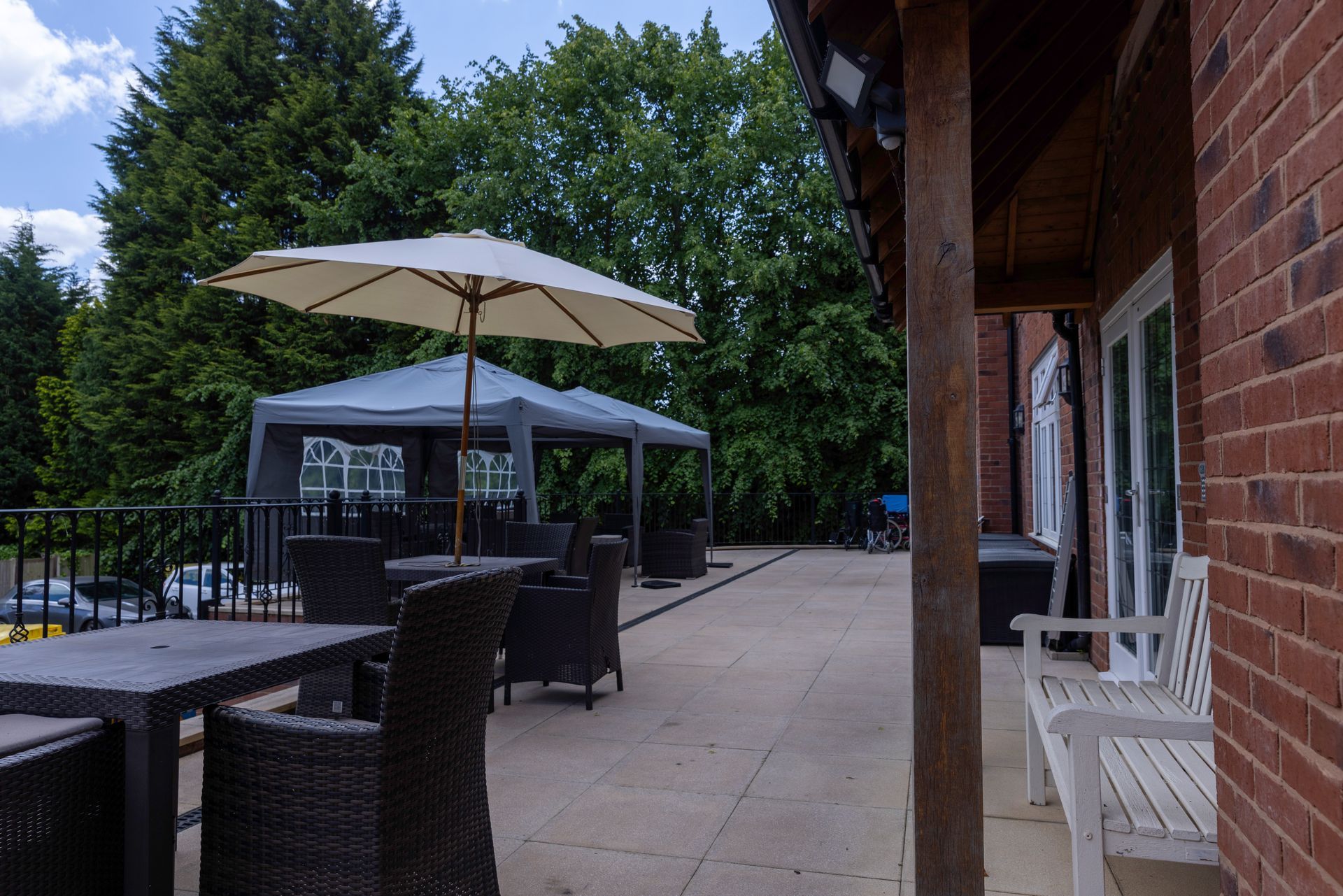 Outdoor patio with tables, chairs, and a parasol under a canopy, next to a brick building with greenery in the background.