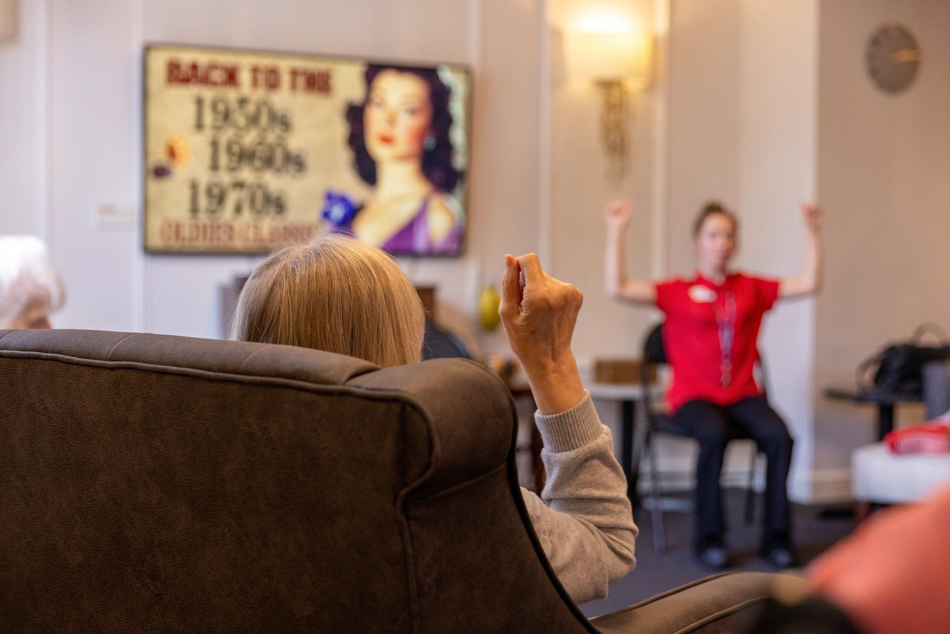 Elderly person in a chair raises a fist during a seated exercise class, led by an instructor in a red shirt.